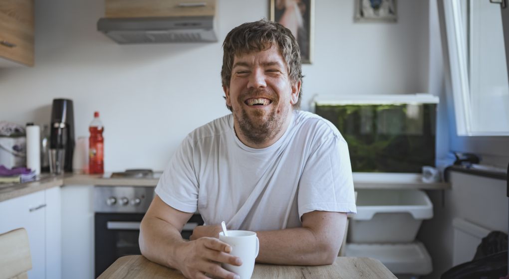 Ein lachender Mann in einem weißen T-Shirt sitzt an einem Küchentisch und hält eine Tasse. Im Hintergrund sind Küchenutensilien und ein Aquarium zu sehen.